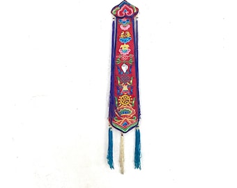 Könnte beinhalten: Ein langer, vertikaler Wandbehang aus rotem Stoff mit komplexer, mehrfarbiger Stickerei verschiedener Symbole, darunter ein Rad und Lotusblumen. Das Stück ist mit langen violetten Fransen gesäumt und endet in drei herabhängenden Quasten in Petrol und Creme.