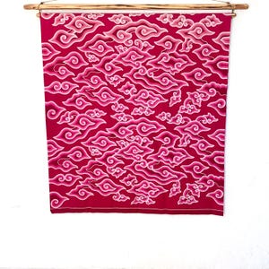 以下が含まれることがあります： ピンクと白の色合いの雲模様が特徴の赤い布製の壁掛け。布は上部の木の棒から吊り下げられています。雲のデザインは、布の表面全体に繰り返されています。