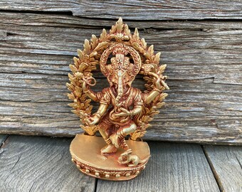 May include: Gold-colored metal statue of the Hindu deity Ganesha, depicted in a dancing pose with four arms. The statue is surrounded by a decorative floral design and stands on a circular base.