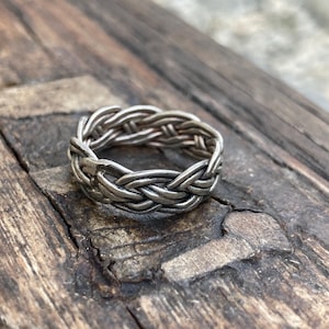 May include: A silver braided ring with a woven design. The ring is lying on a wooden surface.