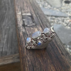 K&ouml;nnte beinhalten: Ein silberner Ring mit mehreren Mondstein-Edelsteinen in verschiedenen Formen und Gr&ouml;&szlig;en. Der Ring hat ein detailliertes Blumendesign um die Steine herum und einen Halbmond-Akzent. Der Ring wird auf einer verwitterten Holzoberfl&auml;che pr&auml;sentiert.