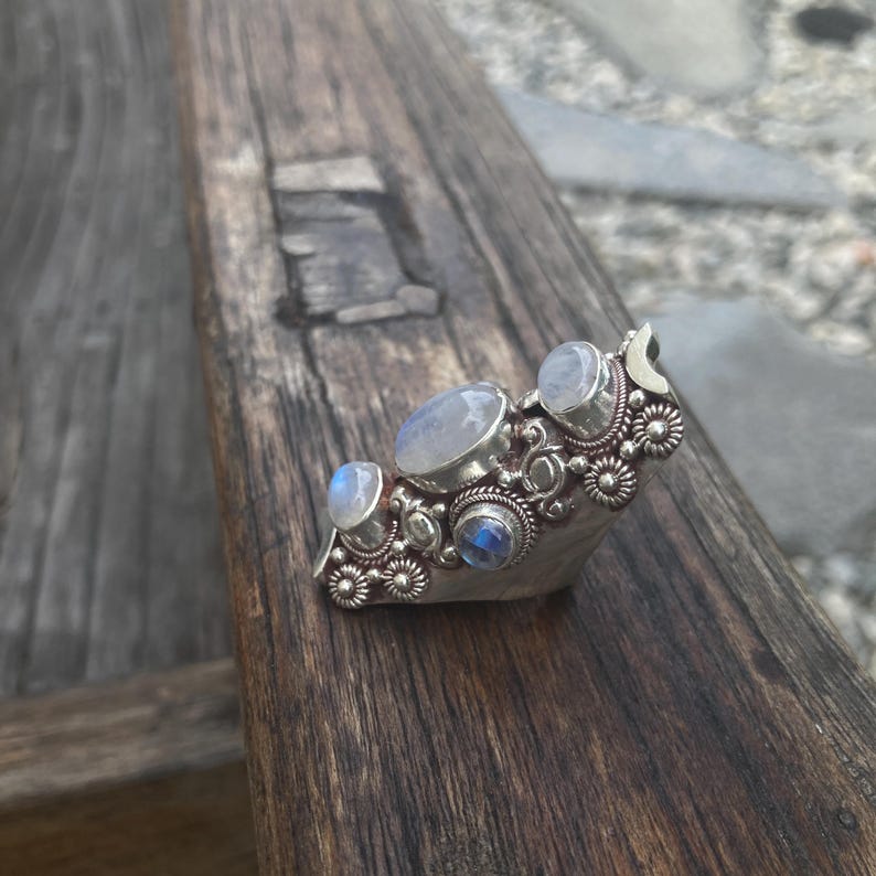K&ouml;nnte beinhalten: Ein silberner Ring mit mehreren Mondstein-Edelsteinen in verschiedenen Formen und Gr&ouml;&szlig;en. Der Ring hat ein detailliertes Blumendesign um die Steine herum und einen Halbmond-Akzent. Der Ring wird auf einer verwitterten Holzoberfl&auml;che pr&auml;sentiert.