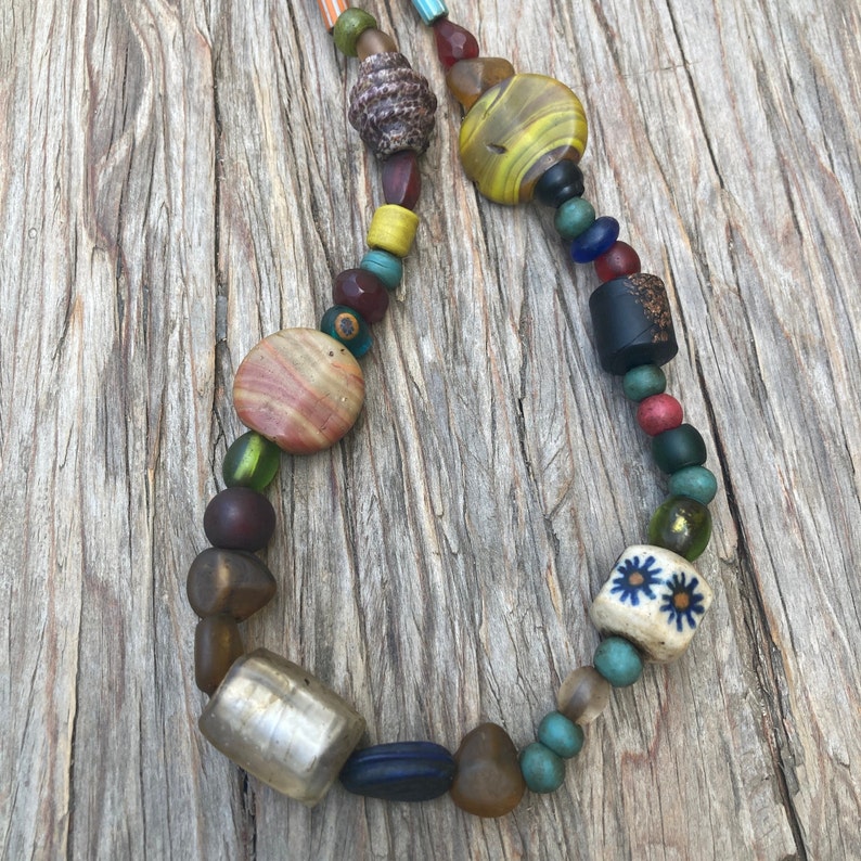 May include: A colourful beaded necklace with a variety of glass, wood, and metal beads. The necklace features a mix of colours, including blue, green, red, yellow, and brown. The beads are strung on a clear cord.