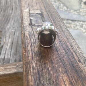 K&ouml;nnte beinhalten: Ein silberner Ring mit einem gro&szlig;en, klaren, oval geformten Stein in der Mitte, flankiert von zwei kleineren, runden, blau-grauen Steinen. Der Ring liegt auf einer Holzoberfl&auml;che und zeigt sein aufwendiges Design und seine Handwerkskunst. Der Ringreif ist sichtbar.