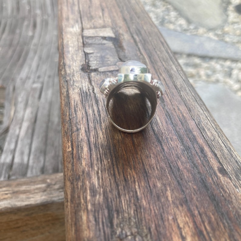 K&ouml;nnte beinhalten: Ein silberner Ring mit einem gro&szlig;en, klaren, oval geformten Stein in der Mitte, flankiert von zwei kleineren, runden, blau-grauen Steinen. Der Ring liegt auf einer Holzoberfl&auml;che und zeigt sein aufwendiges Design und seine Handwerkskunst. Der Ringreif ist sichtbar.