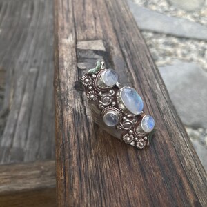 K&ouml;nnte beinhalten: Ein silberner Ring mit mehreren Mondstein-Cabochons. Der Ring hat ein verziertes Design mit floralen und kreisf&ouml;rmigen Akzenten. Die Mondsteine sind oval und rund, mit einem blassblauen Farbton. Der Ring liegt auf einer Holzoberfl&auml;che.