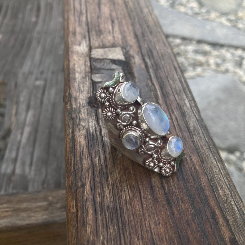 K&ouml;nnte beinhalten: Ein silberner Ring mit mehreren Mondstein-Cabochons. Der Ring hat ein verziertes Design mit floralen und kreisf&ouml;rmigen Akzenten. Die Mondsteine sind oval und rund, mit einem blassblauen Farbton. Der Ring liegt auf einer Holzoberfl&auml;che.
