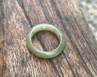 May include: A round, pale green jade ring. The ring is a solid band with a smooth, polished surface. The jade has subtle variations in color, with areas of lighter and darker green. The ring is set against a rustic wooden background.