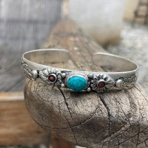 May include: Silver cuff bracelet with a turquoise stone and two red stones. The bracelet has a floral design.