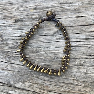 Op de afbeelding: Een donkerbruine koordarmband met een reeks kleine, bungelende druppelvormige bedels van messingkleurig metaal langs de band, afgewerkt met een enkele ronde messingkleurige metalen kraal als sluiting aan de bovenkant van de lus.