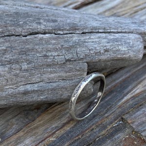May include: A silver ring with an etched pattern of stylized eye shapes. The ring is set against a weathered, gray wooden surface, creating a rustic aesthetic. The ring's design is repeated around the band.