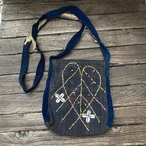 May include: A dark gray fabric shoulder bag with a long blue strap featuring cream-colored lace trim. The front is decorated with a lattice pattern of multicolored seed beads, small metallic charms, and two white cowrie shell flower accents.