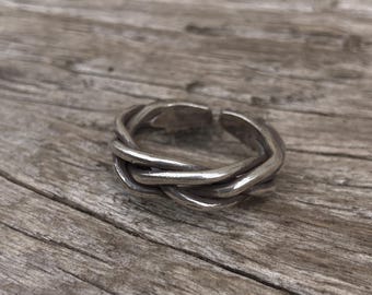 Könnte beinhalten: Ein silberfarbener Ring mit geflochtenem Design. Der Ring scheint aus Metall zu sein und hat eine strukturierte Oberfläche. Der Ring wird auf einer Holzoberfläche präsentiert.