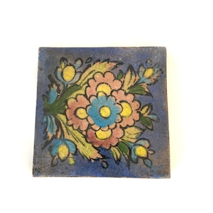 May include: A blue ceramic tile with a floral design. The flowers are pink, yellow, and blue with black outlines. The tile has a distressed look.