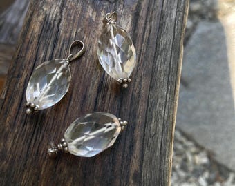 May include: Three clear, faceted crystal pendants with silver-coloured metal findings. The pendants are oval-shaped and have a geometric design. They are displayed on a weathered, brown wooden surface.