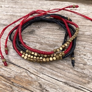 May include: A set of three bracelets with red and black cords and gold-toned metal beads. The bracelets are tied together with a knot.