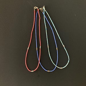 May include: Three beaded necklaces in shades of coral, blue, and turquoise. Each necklace features small, colorful beads strung together with silver-toned accents and clasps. The necklaces are displayed against a dark background.