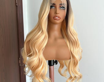 May include: A long, wavy blonde wig with dark roots and a lace front is displayed on a mannequin head. The wig features a blend of blonde and light brown tones, creating a natural, sun-kissed appearance. The hair is styled in loose, flowing waves.