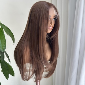 Puede incluir: Una peluca marrón con encaje frontal, con capas largas y rectas, se muestra en una cabeza de maniquí. La peluca tiene una línea de cabello de aspecto natural y un color marrón medio. El cabello cae hasta los hombros. El maniquí está sobre un fondo blanco.