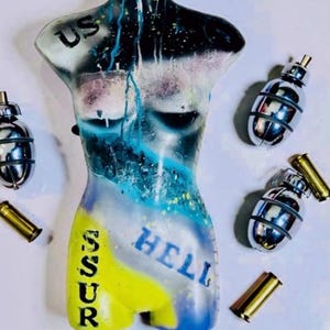 May include: A torso mannequin painted with blue, black, and yellow spray paint, featuring the text US, SSUR, and HELL. The mannequin is surrounded by four metallic silver hand grenades and three brass-coloured bullet casings on a white surface.