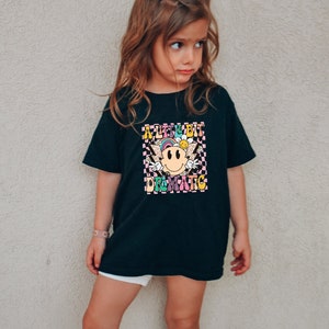 May include: A black t-shirt with a colorful graphic print featuring a smiley face with wings and the text "A Little Bit Dramatic".