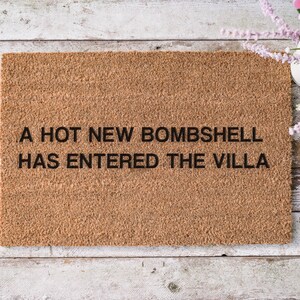May include: A rectangular doormat made of natural brown coir fiber. The mat features the phrase "A HOT NEW BOMBSHELL HAS ENTERED THE VILLA" in black block letters. The mat is placed on a light-colored wooden surface.