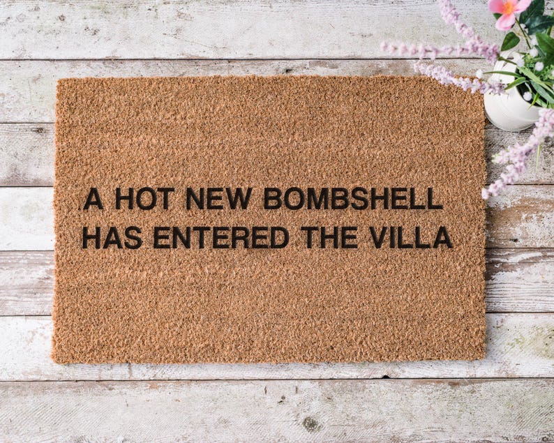 May include: A rectangular doormat made of natural brown coir fiber. The mat features the phrase "A HOT NEW BOMBSHELL HAS ENTERED THE VILLA" in black block letters. The mat is placed on a light-colored wooden surface.