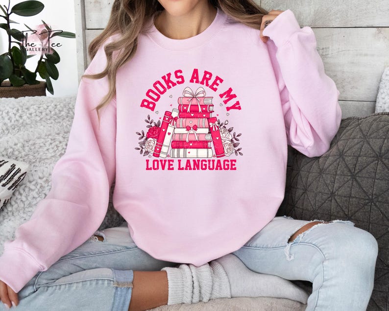 Model posing with one leg tucked under her sporting a pink crewneck sweatshirt with a pink and white design that says books are my love language surrounding a pile of books and a bow on top.