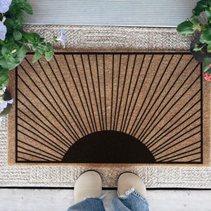 May include: A rectangular doormat with a sunburst design in black on a natural coir background. The mat is surrounded by a woven border and is placed on a wooden porch. Potted flowers in shades of blue, white, yellow, and pink flank the mat.