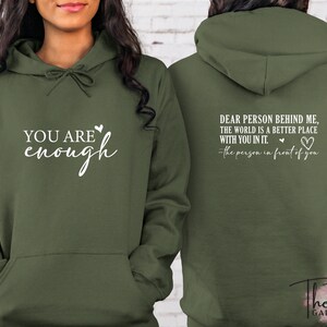 Pu&ograve; includere: Felpa con cappuccio verde oliva con la scritta "YOU ARE enough" sul davanti. Il retro della felpa ha la scritta "DEAR PERSON BEHIND ME, THE WORLD IS A BETTER PLACE WITH YOU IN IT." e "-the person in front of you."
