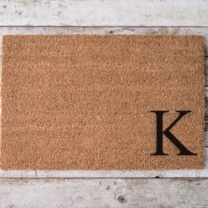 May include: A rectangular doormat made of natural coir fibers. The mat is a light brown color and features a large, black letter "K" in the lower right corner. The doormat is designed for outdoor use.