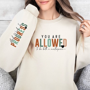 May include: A cream colored sweatshirt with the text "And Work In Progress" on one sleeve and "You Are Allowed To Be Both A Masterpiece..." on the front. The text is in a colorful, layered font with a black heart.