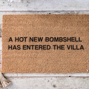 May include: A rectangular coir doormat with the text "A HOT NEW BOMBSHELL HAS ENTERED THE VILLA" in black. The mat is a natural brown color and is placed on a light-colored wooden surface.