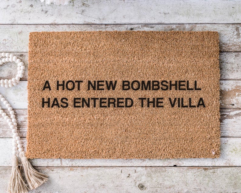 May include: A rectangular coir doormat with the text "A HOT NEW BOMBSHELL HAS ENTERED THE VILLA" in black. The mat is a natural brown color and is placed on a light-colored wooden surface.