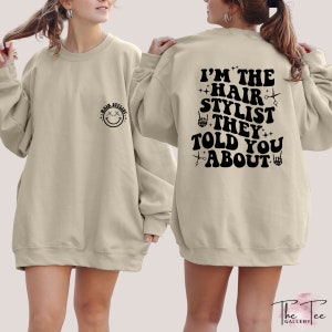 May include: A tan sweatshirt with a black smiley face on the left side and the text "I'M THE HAIR STYLIST THEY TOLD YOU ABOUT" on the back. The text is in a retro font with scissors and hand symbols.