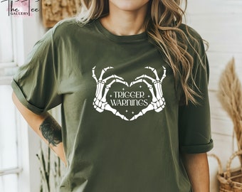 Op de afbeelding: Een groen T-shirt met een witte afbeelding van twee skelethandjes die een hart vormen met de tekst "TRIGGER WARNINGS" erin.