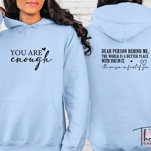 Pu&ograve; includere: Felpa con cappuccio azzurra con la scritta "YOU ARE enough" sul davanti. Il retro della felpa ha la scritta "DEAR PERSON BEHIND ME, THE WORLD IS A BETTER PLACE WITH YOU IN IT." con un simbolo a forma di cuore. Il logo The Tee Gallery &egrave; in basso.