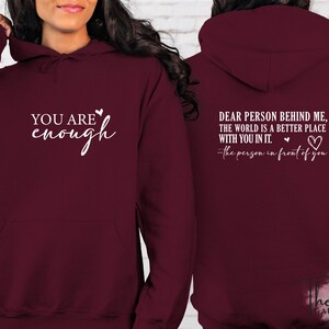 Pu&ograve; includere: Una felpa con cappuccio bordeaux. La parte anteriore mostra il testo "YOU ARE enough" in bianco, con un simbolo a cuore. Il retro della felpa con cappuccio presenta il testo "DEAR PERSON BEHIND ME, THE WORLD IS A BETTER PLACE WITH YOU IN IT." e un simbolo a cuore.