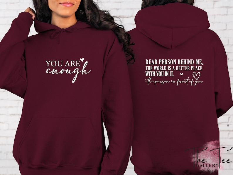 Pu&ograve; includere: Una felpa con cappuccio bordeaux. La parte anteriore mostra il testo "YOU ARE enough" in bianco, con un simbolo a cuore. Il retro della felpa con cappuccio presenta il testo "DEAR PERSON BEHIND ME, THE WORLD IS A BETTER PLACE WITH YOU IN IT." e un simbolo a cuore.