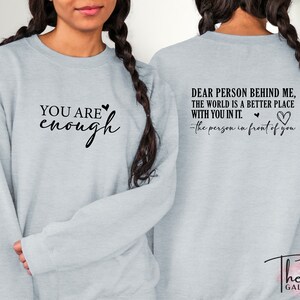 Pu&ograve; includere: Felpa grigio chiaro con la scritta "YOU ARE enough" sul davanti e "DEAR PERSON BEHIND ME, THE WORLD IS A BETTER PLACE WITH YOU IN IT. - the person in front of you" sul retro. Il logo The Tee Gallery &egrave; in basso.