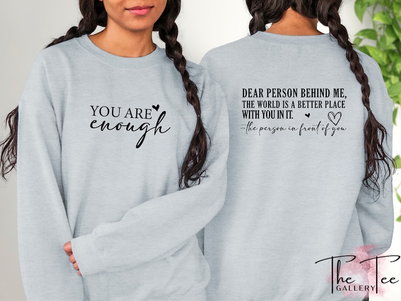 Pu&ograve; includere: Felpa grigio chiaro con la scritta "YOU ARE enough" sul davanti e "DEAR PERSON BEHIND ME, THE WORLD IS A BETTER PLACE WITH YOU IN IT. - the person in front of you" sul retro. Il logo The Tee Gallery &egrave; in basso.
