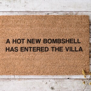 May include: A rectangular doormat made of natural coir fiber. The mat features the text "A HOT NEW BOMBSHELL HAS ENTERED THE VILLA" in black, centered on the mat. The mat is placed on a light-colored wooden surface.