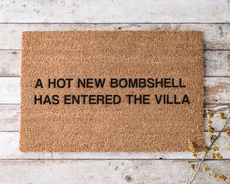 May include: A rectangular doormat made of natural coir fiber. The mat features the text "A HOT NEW BOMBSHELL HAS ENTERED THE VILLA" in black, centered on the mat. The mat is placed on a light-colored wooden surface.
