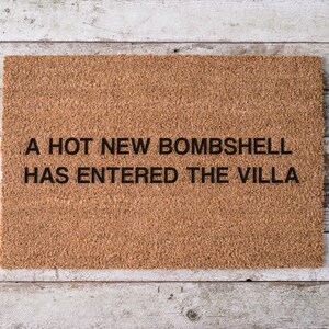 May include: A rectangular doormat made of natural brown coir fiber. The mat features black text that reads "A HOT NEW BOMBSHELL HAS ENTERED THE VILLA". The doormat is placed on a light-colored wooden surface.
