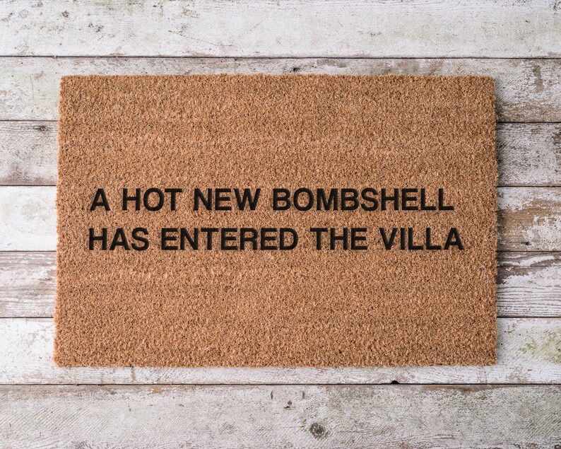 May include: A rectangular doormat made of natural brown coir fiber. The mat features black text that reads "A HOT NEW BOMBSHELL HAS ENTERED THE VILLA". The doormat is placed on a light-colored wooden surface.