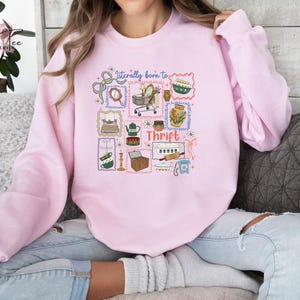 May include: Pale pink sweatshirt featuring a colourful graphic design. The design includes the words "Literally born to Thrift" and illustrations of thrift store items, including a mirror, teapot, and shopping cart.