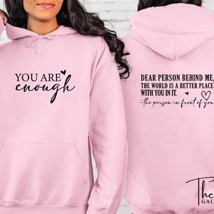 Pu&ograve; includere: Una felpa con cappuccio rosa chiaro con la scritta "YOU ARE enough" sul davanti e "DEAR PERSON BEHIND ME, THE WORLD IS A BETTER PLACE WITH YOU IN IT. -the person in front of you" sul retro. Ha un cappuccio con coulisse e una tasca frontale.