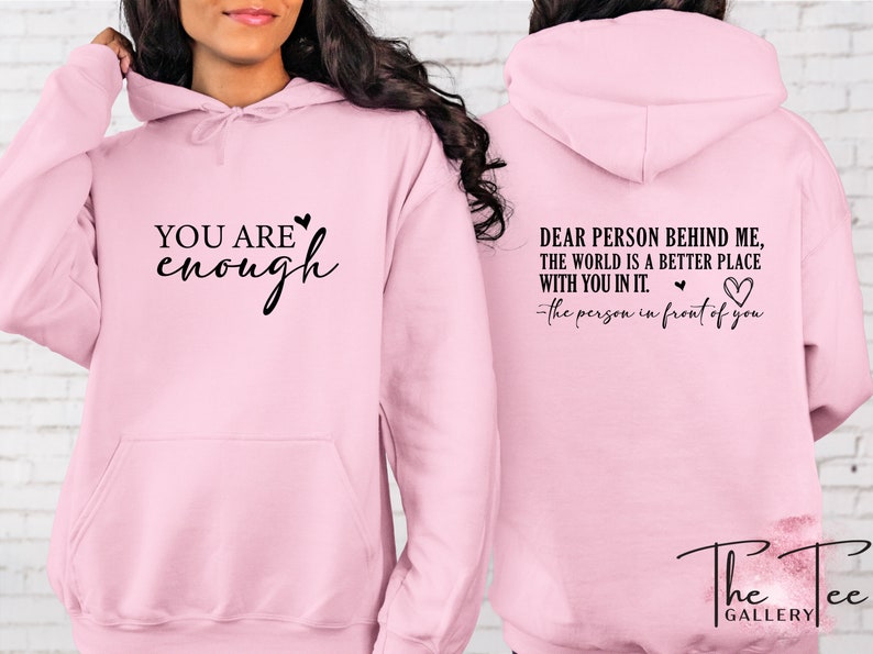 Pu&ograve; includere: Una felpa con cappuccio rosa chiaro con la scritta "YOU ARE enough" sul davanti e "DEAR PERSON BEHIND ME, THE WORLD IS A BETTER PLACE WITH YOU IN IT. -the person in front of you" sul retro. Ha un cappuccio con coulisse e una tasca frontale.