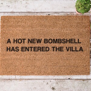 May include: A rectangular doormat made of natural coir fiber. The mat features the phrase "A HOT NEW BOMBSHELL HAS ENTERED THE VILLA" in black block letters. The mat is placed on a light-colored wooden surface.