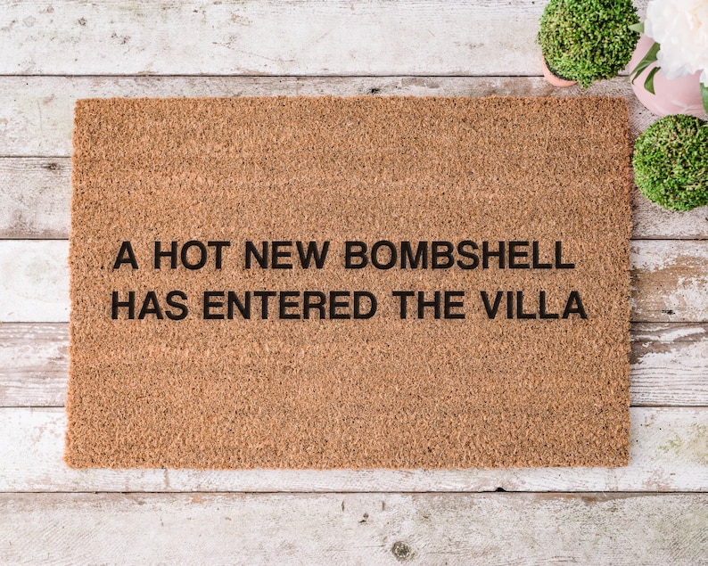 May include: A rectangular doormat made of natural coir fiber. The mat features the phrase "A HOT NEW BOMBSHELL HAS ENTERED THE VILLA" in black block letters. The mat is placed on a light-colored wooden surface.